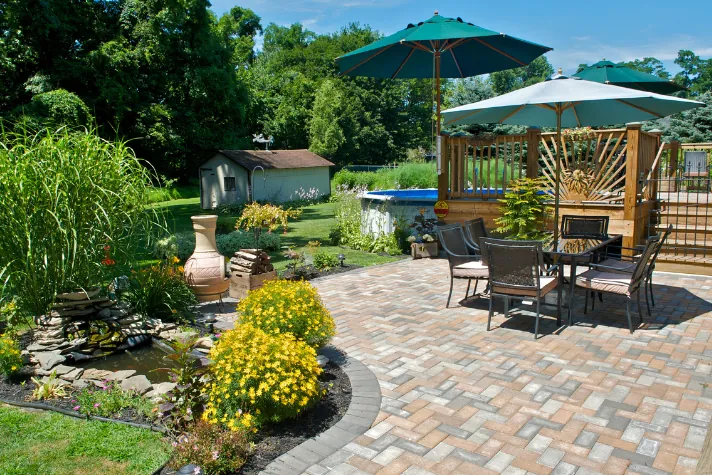 A paver patio with a dining table under green umbrellas beside a wooden pergola and planted beds