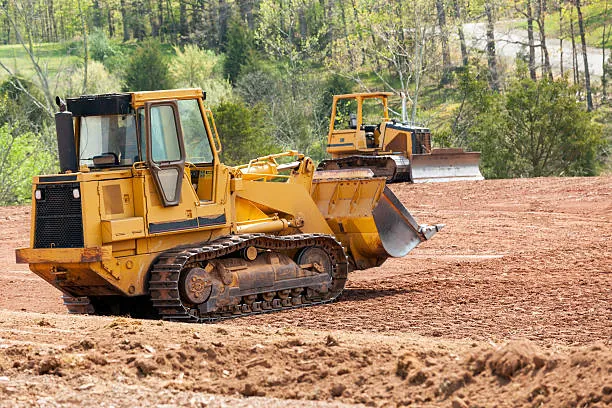 A yellow track loader pushing soil on a cleared lot with trees behind it